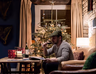 man drinking a glass of wine and reading a menu at a table beside a Christmas tree
