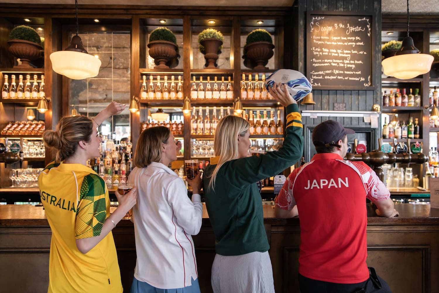 friends wearing rugby jerseys while standing in front of a bar holding a rugby ball and Young's pints